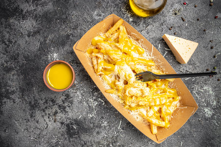 French fries baked with cheddar cheese, pickle and sausages in a paper plate on a dark background,...