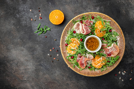 plate of arugula salad with mandarins, prosciutto jamon and Parmesan cheese on a dark background. banner, menu, recipe place for text, top view.の写真素材
