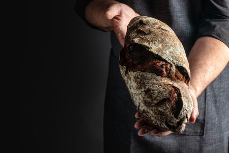 Baker man holding rustic organic loaf of bread in hands on a dark background. banner, menu, recipe place for text, top view,の写真素材