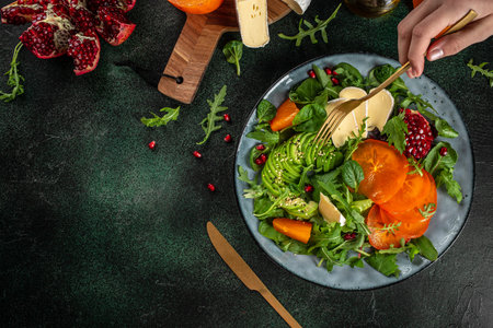 delicious winter salad with persimmon, fresh greens, avocado, Camembert cheese and pomegranate on a dark background. top view. copy space for text,の写真素材