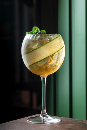 Elegant cocktail with ice, cucumber ribbon, and mint garnish served in a wine glass on a dark wooden...