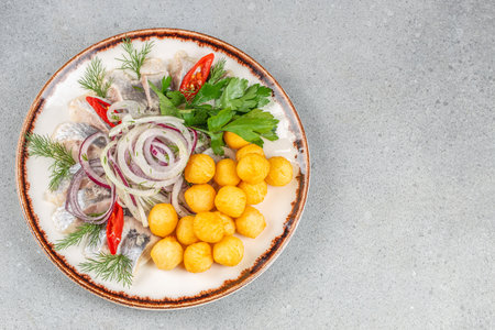 A Nordic-inspired appetizer featuring pickled herring fillets garnished with onion rings, fresh herbs, spicy chili, and golden potato balls on a rustic plate.の写真素材