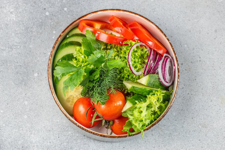 A fresh and colorful vegetable salad with cucumbers, tomatoes, red onions, bell peppers, lettuce, and herbs arranged in a rustic bowl on a gray stone surface.の写真素材