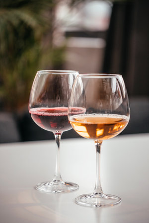 Two elegant wine glasses filled with red and amber wine stand on a white surface, reflecting soft...