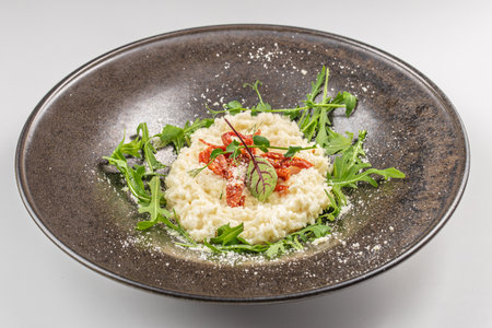 A creamy risotto garnished with sun-dried tomatoes, parmesan cheese, and fresh arugula, beautifully presented on a dark ceramic plate.の写真素材