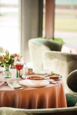 Elegant restaurant table setting with a bowl of hot soup, wine glass, and floral decoration in warm...