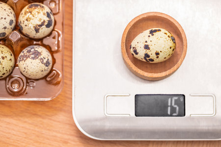 A speckled quail egg is weighed on a digital kitchen scale, displaying a weight of 15 grams beside a plastic egg tray on a wooden surface.の写真素材