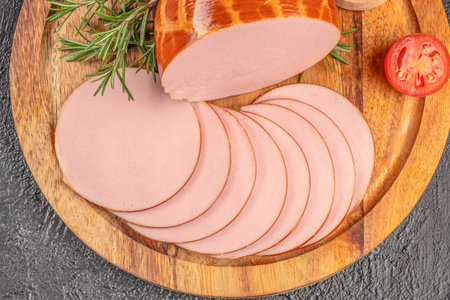 Sliced bologna sausage arranged on a wooden board with rosemary and tomato on a dark background.の写真素材