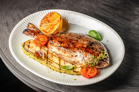 Grilled whole fish served with charred zucchini, roasted tomatoes, and a caramelized lemon on a...