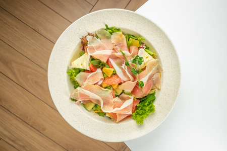 Fresh salad with prosciutto, citrus fruits, cheese, and lettuce served in a white bowl.の写真素材