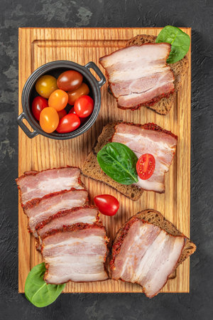Smoked slices on bread with cherry tomatoes and spinach leaves on a wooden board with copy space on dark background.の写真素材
