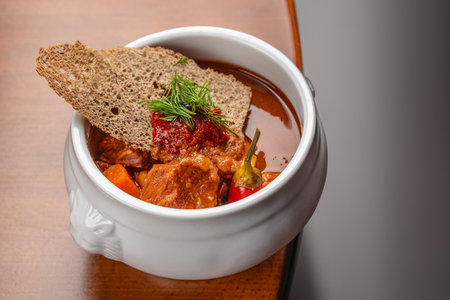 Hot meat stew with vegetables, bread, and spices in a white ceramic bowl on a wooden table with copy space.の写真素材