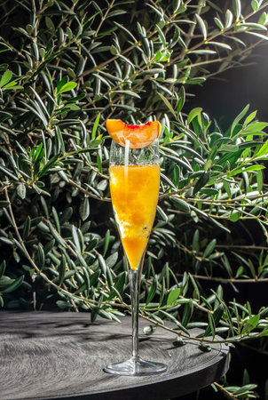 Elegant peach cocktail served in a tall glass with fresh peach garnish, placed on a wooden table against a background of green leaves.の写真素材