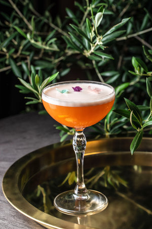 Elegant orange cocktail with foam and edible flower petals, served in a crystal coupe glass on a golden tray, surrounded by green leaves.の写真素材