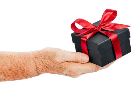 Close-up of elderly hands holding a small black gift box decorated with a red satin ribbon symbol of giving, gratitude, and care, isolated on white background.の写真素材