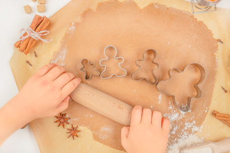 Child rolling gingerbread dough with cookie cutters shaped like gingerbread men, top view, festive Christmas baking scene on parchment paper.の写真素材
