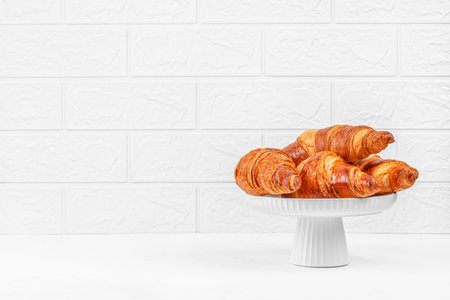 Golden flaky croissants on elegant white cake stand against light brick wall background, bakery pastry display with large copy spaceの写真素材