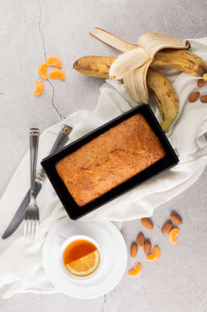 Banana bread, bananas, a cup of tea, a fork, a knife, nuts and tangerine slices on a gray background.の写真素材