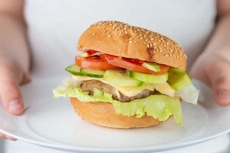 Blurred image of a hamburger with cutlet, cheese and vegetables in a white plate in the hands of a...