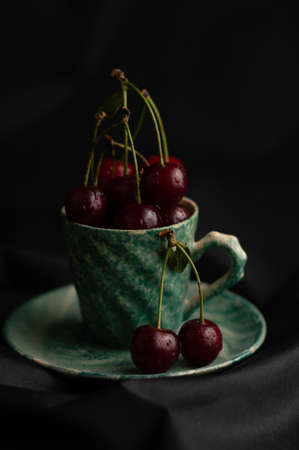 Blurred image of fresh, ripe cherries in a green cup and saucer against a black background.の写真素材