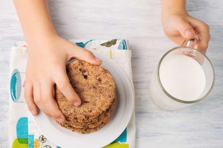 A child takes a giant chocolate chip cookie from a plate on a light table with a cup of milk.の写真素材