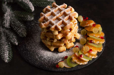 Belgian waffles with powdered sugar and slices of fruit on a dark background with sprigs of spruce. Concept background for Christmas menu or recipe.の写真素材