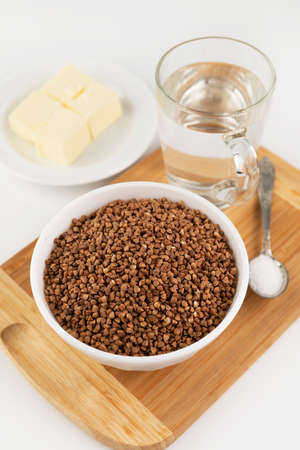 Raw buckwheat in a plate, a glass of water, oil and salt on a cutting board. Ingredients for cooking. Healthy, simple food.の写真素材