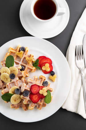 Viennese waffles with berries on a white plate and a cup of coffee on a black table.High-calorie...