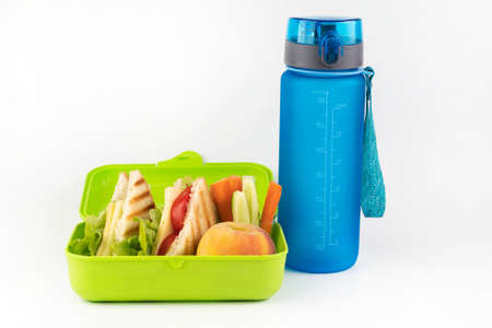 On a white background, a bottle of water, a lunch box with toasts, vegetables and peach. School...