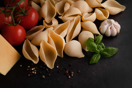 On a dark background, a blurred image of conchiglioni pasta, tomatoes, garlic, basil sprig and cheese.の写真素材