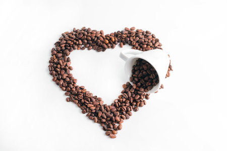 A heart made of coffee beans and a coffee cup is laid out on a white background. Coffee love concept.の写真素材
