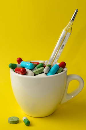 Colorful different pills and medical thermometer inside a cup on yellow background