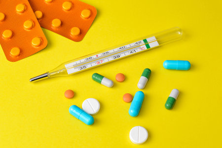 Colorful different pills and medical thermometer on yellow surface