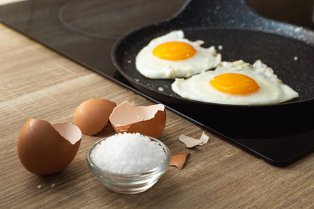 fried eggs with spatula in a frying pan, salt and eggshell on the tableの写真素材