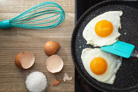 Flat lay of fried eggs with spatula in a frying pan, salt and eggshell on the table.の写真素材
