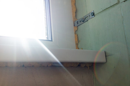 A sunbeam highlights a metal bracket securing a window frame, with insulation foam sealing the space...