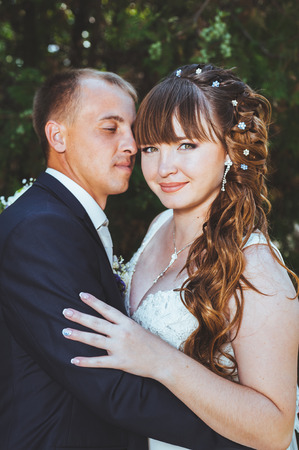 wedding couple kissing in green summer park. bride and groom kissing, standing together outdoors, hugging among green trees. Bride holding wedding bouquet of flowers.の写真素材