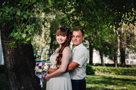 wedding couple kissing in green summer park. bride and groom kissing, standing together outdoors, hugging among green trees. Bride holding wedding bouquet of flowers.の写真素材