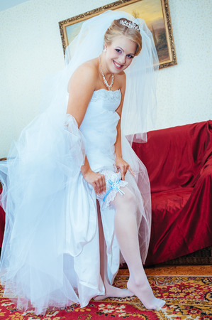 Garter on the leg of a bride, slim sexy bride in wedding luxury dress showing her silk garter with golden ribbon. woman have a final preparation for wedding ceremony. Wedding day moments.の写真素材