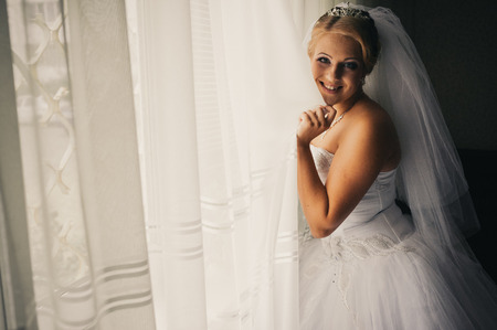 beautiful young bride standing beside a large window waiting. Beautiful caucasian bride getting ready for the wedding ceremonyの写真素材