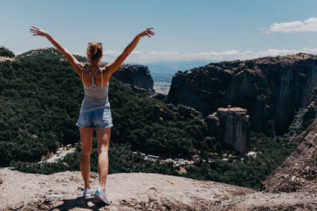 Young happy woman stands back to the camera with raised hands. Traveler enjoying the landscape with mountain, Monasteries of Meteora, Greece.の写真素材