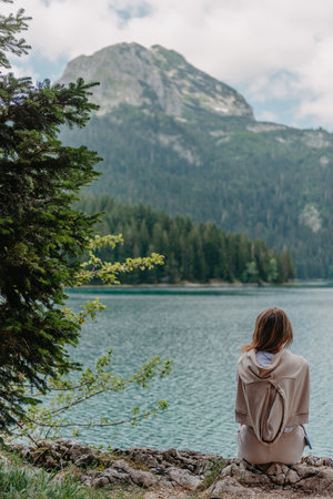 Young woman on shore of mountain lake. The back of girl against background of mountains. Young girl sitting near by beautiful Black Lake in Montenegro. Original wallpaper from summer morningの写真素材