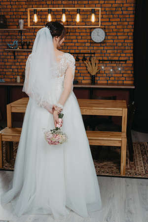 Beautiful bride in long wedding dress and veil with bridal bouquet waiting for groom before wedding ceremony at hotel, back view, copy space. Bride morningの写真素材