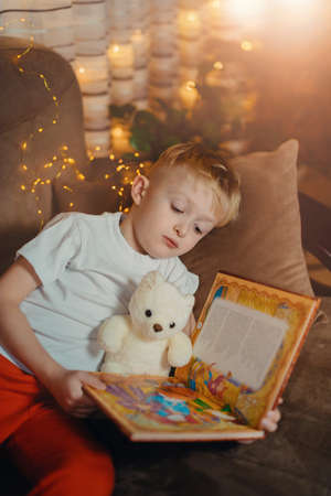 3-4 years old boy is reading a book sitting on a sofa with teddy bear. Home reading of fairy tales. Cute little boy is reading book while sitting on a sofa. Child reads a book to his best bear friend.の写真素材