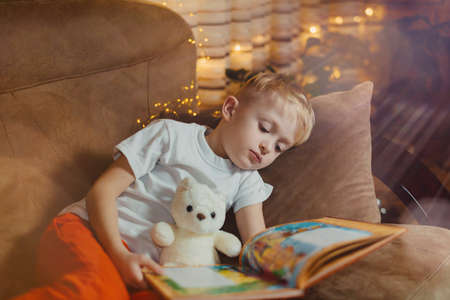 3-4 years old boy is reading a book sitting on a sofa with teddy bear. Home reading of fairy tales. Cute little boy is reading book while sitting on a sofa. Child reads a book to his best bear friend.の写真素材