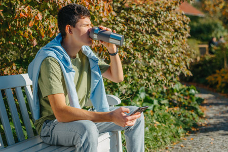 a teenager sits on a bench in the park drinks coffee from a thermo mug and looks into a phone....