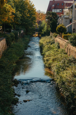 Beautiful small river with clean and clear water front of colorful autumn trees and small old town on the hill agaist nice blue and clouds sky during autumn in Europeの写真素材