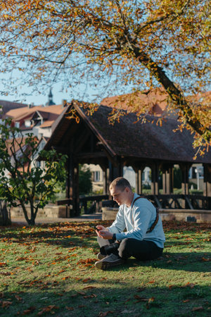 Professional Photographer Taking Picture Of Beautiful Autumn Park. Man Professional Photographer Sit With Camera And With Smartphone In Autumn Park. Retouched, Vibrant Colors, Brownish Tones.の写真素材