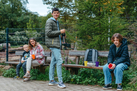 Happy Family drinking tea from a thermos in the forest. Photo of cute charming mother with childrens dressed casual outfit walking sitting bench drinking tea smiling outside city parkの写真素材
