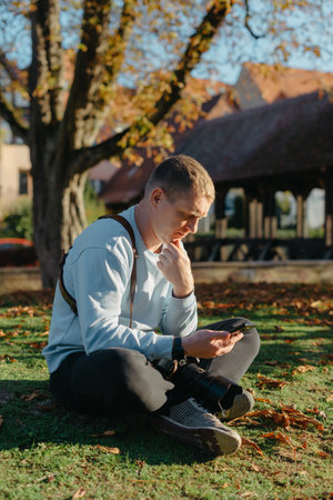 Professional Photographer Taking Picture Of Beautiful Autumn Park. Man Professional Photographer Sit With Camera And With Smartphone In Autumn Park. Retouched, Vibrant Colors, Brownish Tones.の写真素材